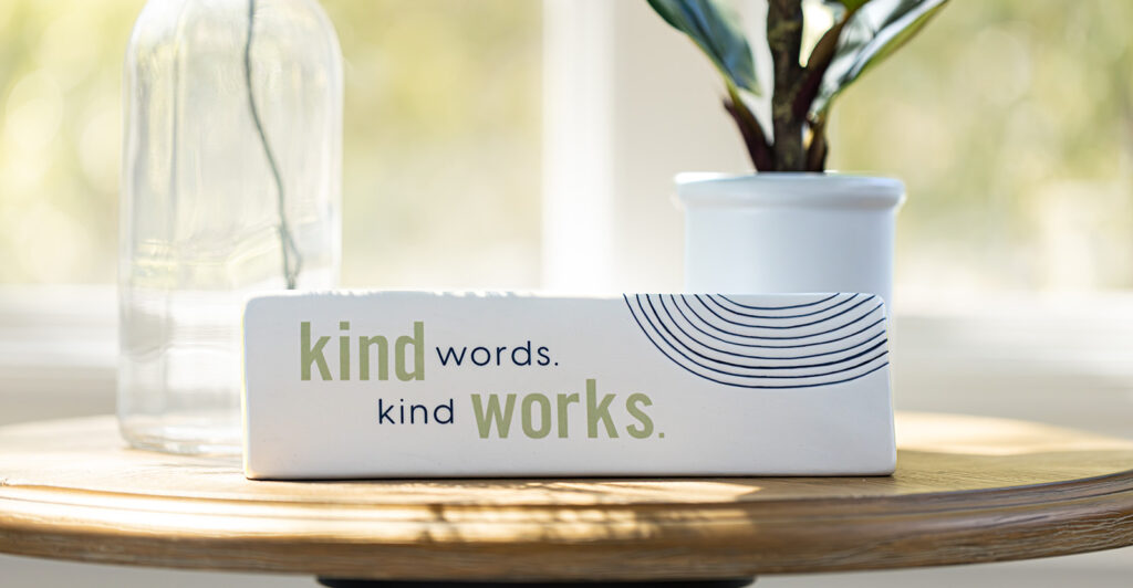 Kind Words Kind Works desk decoration on table in front of plant and vase.