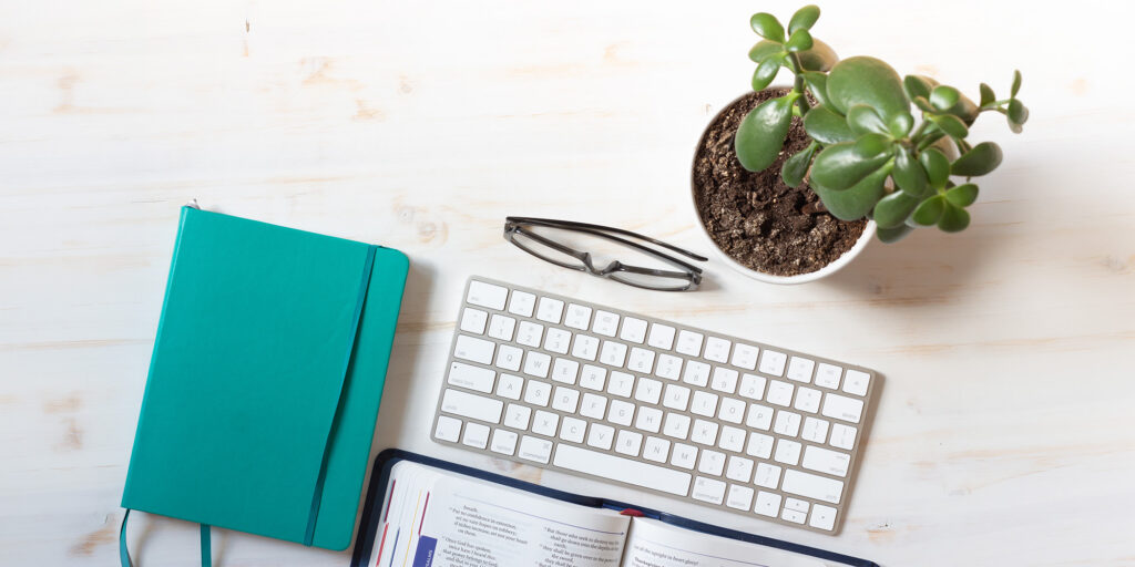 Tabletop view of desk with journal, keyboard, glasses and plant.