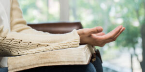 A woman sitting on a couch with the Bible in her lap and open hands resting on top.